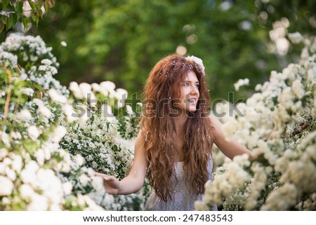 Beautiful lady in spring garden full of white flowers. She touches branches and flowers by hands. young woman in white dress with long brunette curly hear and big white flower in her hear. Look aside