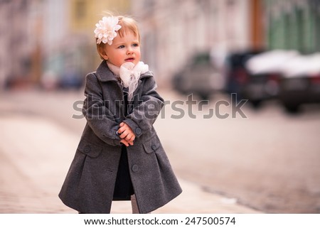 portrait of little baby in gray coat on the street in old city with funny smile. One year old girl dressed on grey coat with big white flower in hear and white bow around neck. She puts hands together