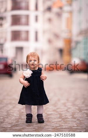 little lady stand on the street. One year old girl dressed on dark velvet dress with big white flower and white tights. The background is diffused with yellow and brown colors