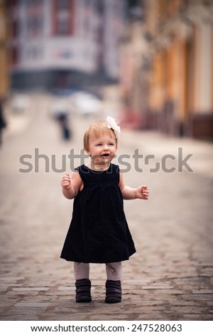 little lady with charming smile stand on the street. One year old girl dressed on dark velvet dress with big white flower and white tights. The background is diffused with yellow and brown colors
