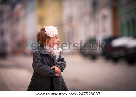 portrait of little baby girl in gray coat on the street in old city with funny smile. One year old girl dressed on grey coat with big white flower in her hear and big white bow around neck. look back
