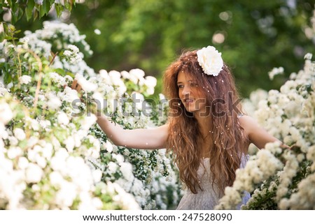 Beautiful lady in spring garden full of white flowers. She touches branches and flowers by hands.Young brunette woman in white dress with long curly hear and big white flower and flower petals in hear