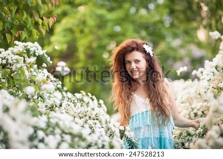 Beautiful lady in spring garden full of white flowers.She touches branches and flowers. Young brunette woman in blue and white dress with long curly hear and big white flower and flower petals in hear