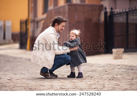 Father takes his little baby in his arms on the street with stone paving. Girl is dressed on grey coat and dress and with white flower in her hair. Young man dressed on white jacket and jeans