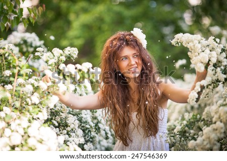 Beautiful brunette lady in spring garden full of white flowers. She touches branches and flowers by hands. Young woman in white dress with long curly hear and big white flower in her hear