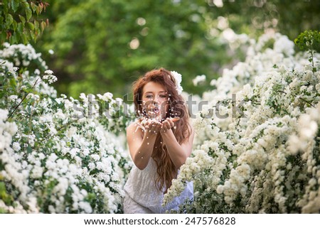 Beautiful brunette lady in spring garden full of white flowers touches branches and flowers by hands.Young woman in white dress with long curly hear and big white flower and petals in hear