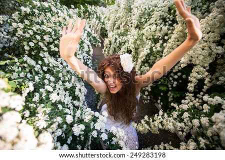 Beautiful brunette lady with bright smile in spring garden full of white flowers. It is young woman in white dress with long curly hear and big white flower and petals in hear