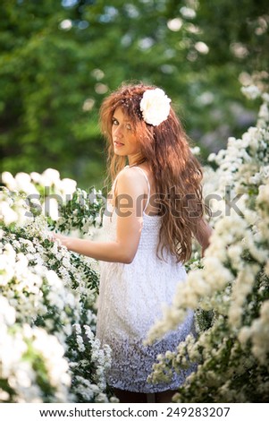 Beautiful brunette lady in spring garden full of white flowers touches branches and flowers. Look back. It is young woman in white dress with long curly hear and big white flower and petals in hear