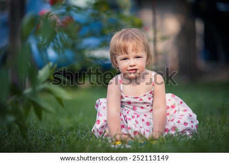 baby girl with blue eyes and blond hair sits on the grass and plays toy. She dressed on white with floral pattern summer dress. Background is diffused by green, brown and blue colors