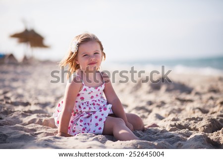 little blond girl with blue eyes on the beach near the sea dressed on white sun-dress with white flower in hair. Background diffused. Small lady play in the sand