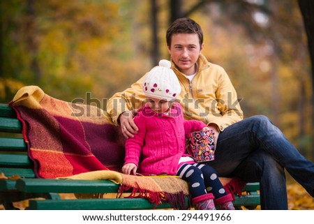 Little girl in bright clothes sits on the bench with her father in autumn garden. Bench covered by plaid. Girl holds colored pail. Father hugs daughter