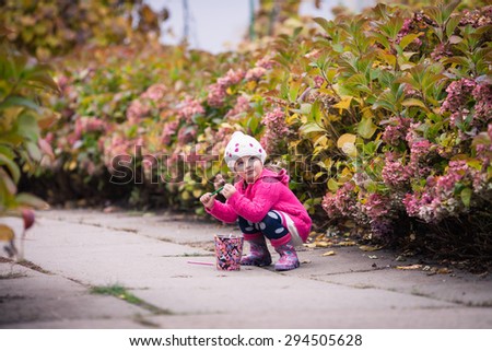 Little girl in bright clothes  went into a crouch in autumn garden. Baby plays with bright colored pail and smile
