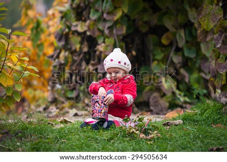 Little girl in bright clothes sits on the grass in autumn garden.  Baby holds colored pencil and bright colored pail