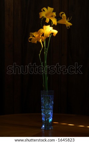 Blue glass vase with glass stones and tall daylilies on oak table, lit with light painting.