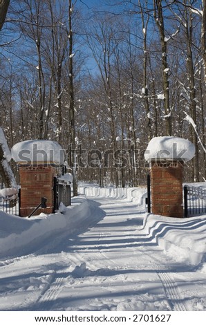 A winter driveway in the country, winding through a snow covered path and brick and iron gateposts, with deciduous forest on either side.