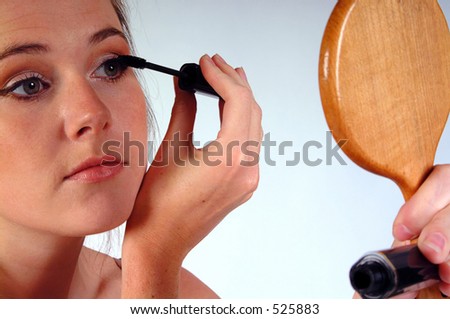 Young woman gazing into a hand mirror whilst applying her mascara. Shot against a white background.