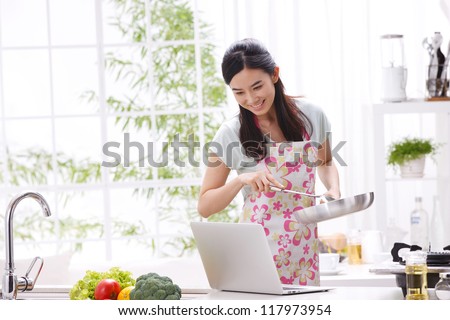 Young woman in kitchen cooking with reference the laptop