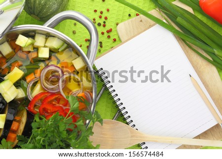 Blank recipe book with vegetable soup, kitchen equipment and veggies around them