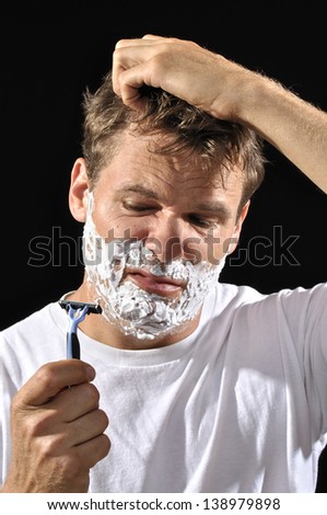 Man with shaving cream on face looks disgruntledly at his razor and scratches his head