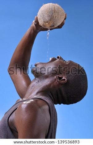 Young lean athletic black man pours fresh pure coconut water directly from fruit to his mouth on blue sky background
