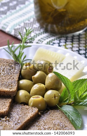 Healthy plate of Mediterranean diet food including stuffed green olives, whole wheat bread, European cheese, mint leaves, rosemary, and olive oil