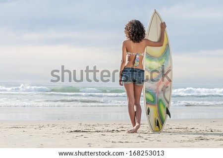 Beautiful young surfer girl standing with surfboard in sand watching beautiful waves in ocean