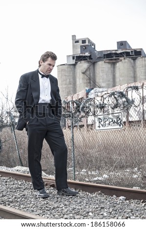 Depressed Caucasian man in formal black tuxedo walks along railroad tracks in urban industrial zone