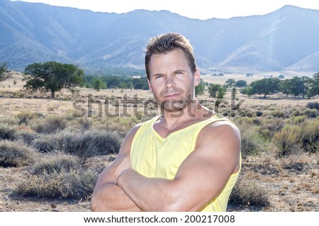 Tough looking muscular Caucasian man with arms crossed looks into camera while standing in natural outdoor countryside surroundings