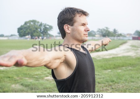 Athletic Caucasian man in black tank top extends out arms while doing warm up stretch at ball field
