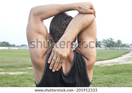 Muscular lean Caucasian man wearing black tank top performs triceps stretch while looking at ball field