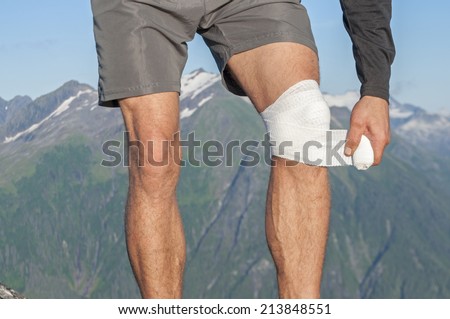 Male runner wearing shorts on top of mountain with beautiful scenic view wraps injured knee with white sports bandage