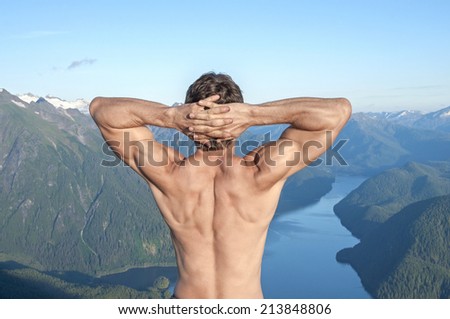 Back of shirtless muscular Caucasian man stretching arms with fingers behind head as he enjoys the magnificent scenic view over body of water surrounded by snow capped mountains near Sitka, Alaska