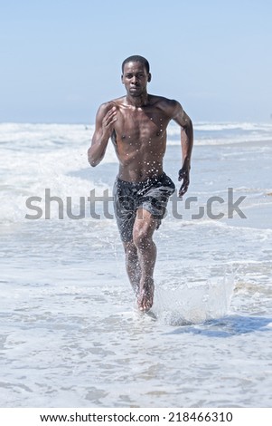 Lean muscular young athletic African American man in shorts runs and splashes through shallow water at beach
