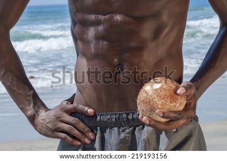 Closeup of lean and fit abdominal muscles of African American man holding whole coconut on beautiful beach