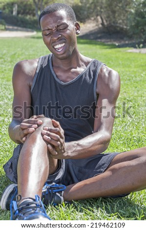 Young lean African American athlete in shorts and tank top sits on grass clutching injured knee and showing painful facial expression