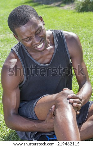 Young lean African American athlete in shorts and tank top sits on grass clutching injured knee and showing painful facial expression