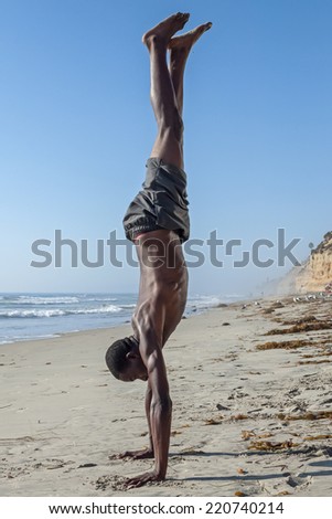 Side view of tall lean shirtless African American man performing handstand on California beach under clear blue sky
