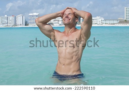 Lean muscular shirtless Caucasian man with abs smiles and pulls hair back while standing in beautiful shallow tropical sea with large resorts in background
