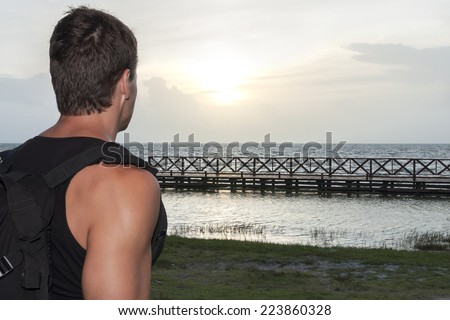 Over the shoulder view of Caucasian male hiker observing the sunrise over a tropical bay with wooden dock in Chetumal, Mexico