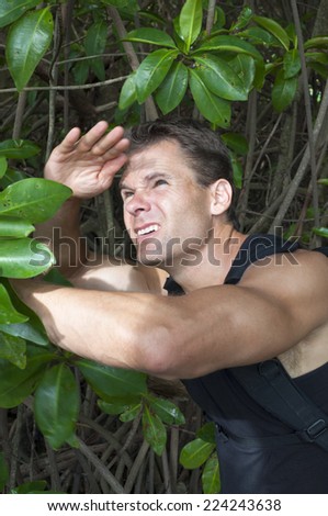 Muscular handsome Caucasian man shields face from bright sun light as he explores thick jungle mangrove swamp