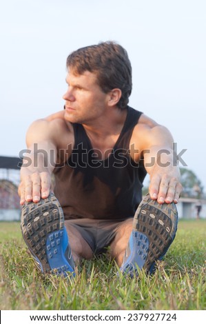 Focus on shoes as muscular Caucasian man sits in grass reaching for toes to do hamstring stretch on grass sport field