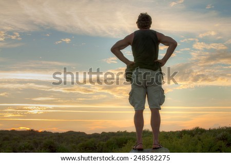 Back of male hiker standing with hands on hips while viewing landscape beneath brilliant sunset in Tulum, Mexico