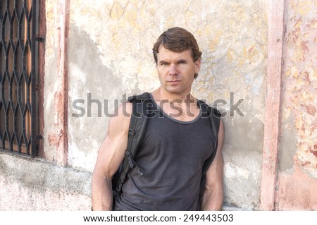 Handsome muscular Caucasian man wearing black tank top and backpack leans against rustic exterior wall in Merida, Mexico