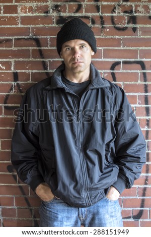 Caucasian man with serious expression wears hoodie, jacket and jeans and leans against brick wall with graffiti in urban setting with hands in pockets