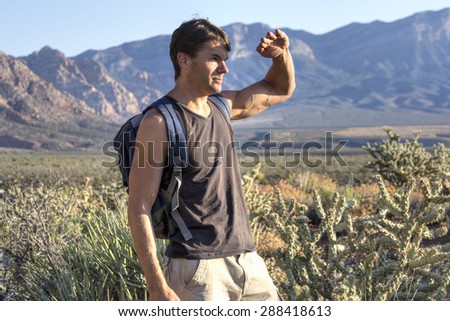 Lean muscular Caucasian man hiking in desert shields eyes from intense morning sunlight