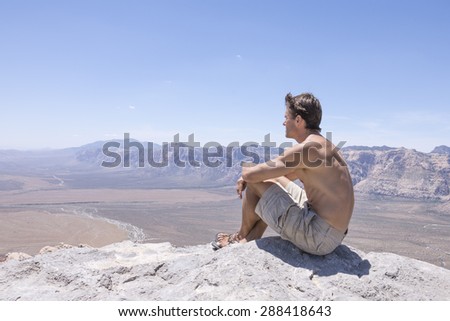 Wide angle of shirtless muscular Caucasian man in shorts and sandals sitting on rocky summit of mountain overlooking expanse of desert under sunny clear sky in Red Rock Canyon, Nevada