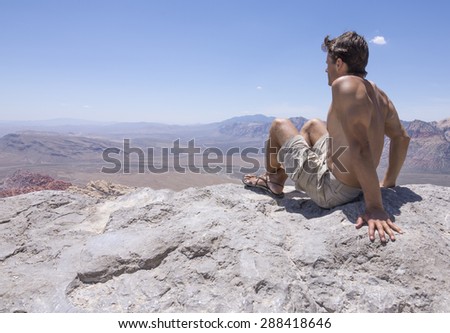 Muscular shirtless Caucasian man wearing shorts and sandals relaxes on top of rocky summit overlooking Red Rock Canyon in Nevada on hot sunny day
