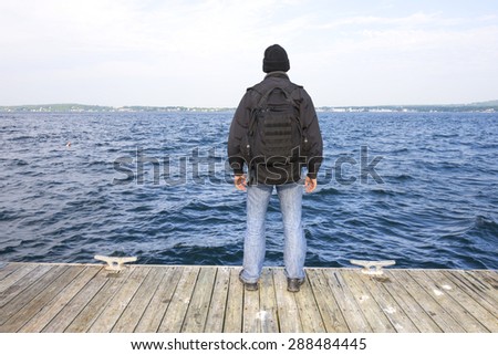 Back side of man with black backpack and jacket standing at end of wooden dock overlooking choppy waters of bay in Rockland, Maine