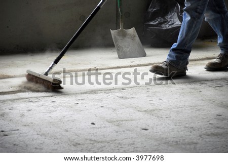 The legs and feet of a construction worker sweeping up on rough concrete at a job site using a large broom