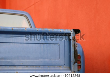 The tailgate of a blue battered sixties American Pickup Truck in front of a red wall